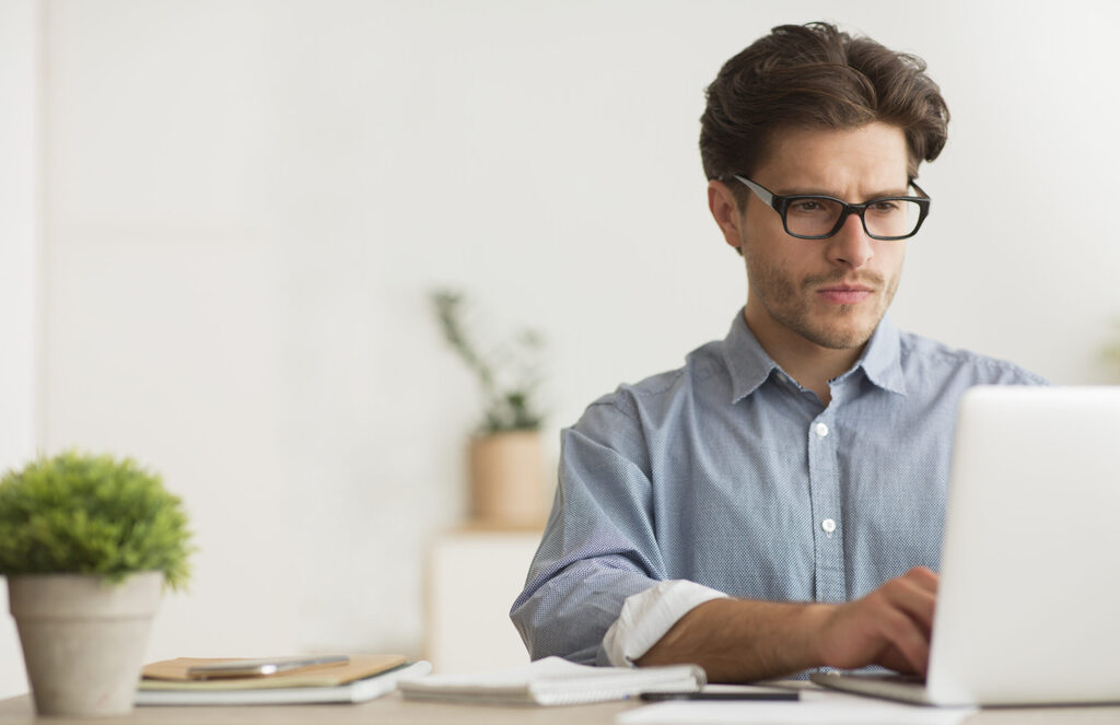 A man wearing glasses is focused on his laptop, engaged in work at a desk.