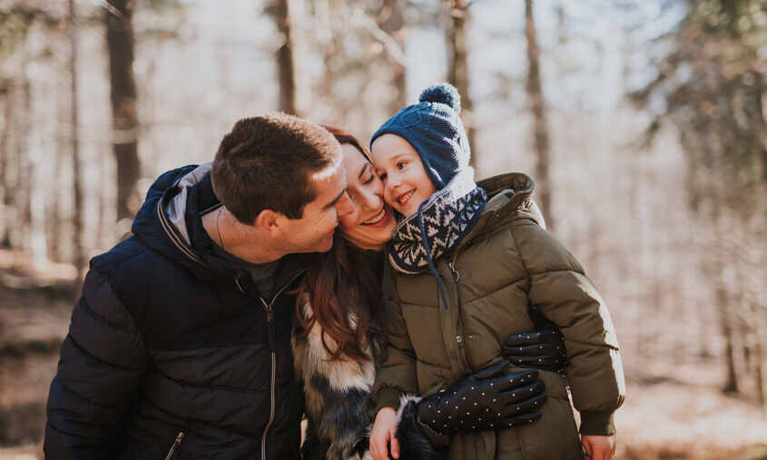 A family of three is outdoors in a forest, dressed in warm clothing. The child, in a winter hat and coat, is smiling while being hugged by the parents.
