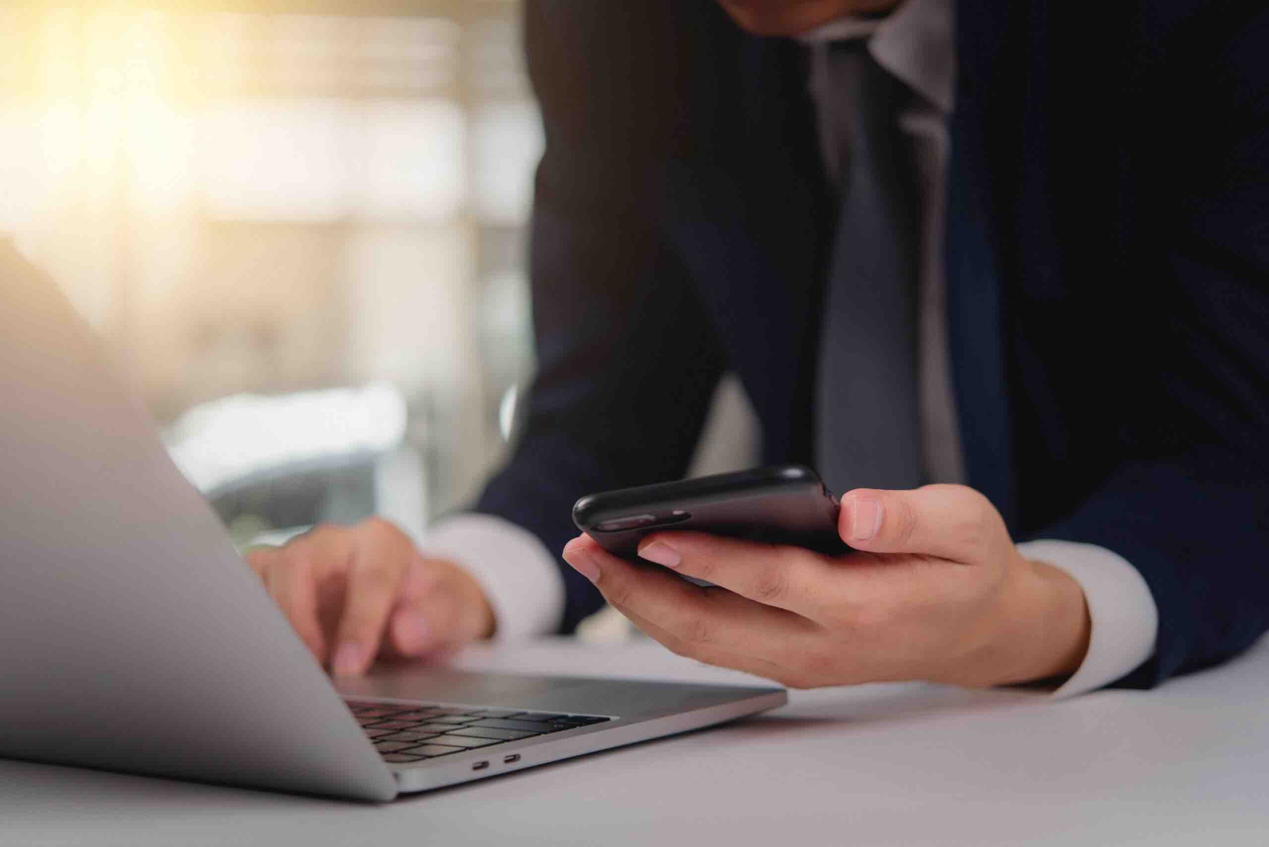 Business professional using a smartphone while working on a laptop. Suitable for modern business communication, remote work, or digital technology