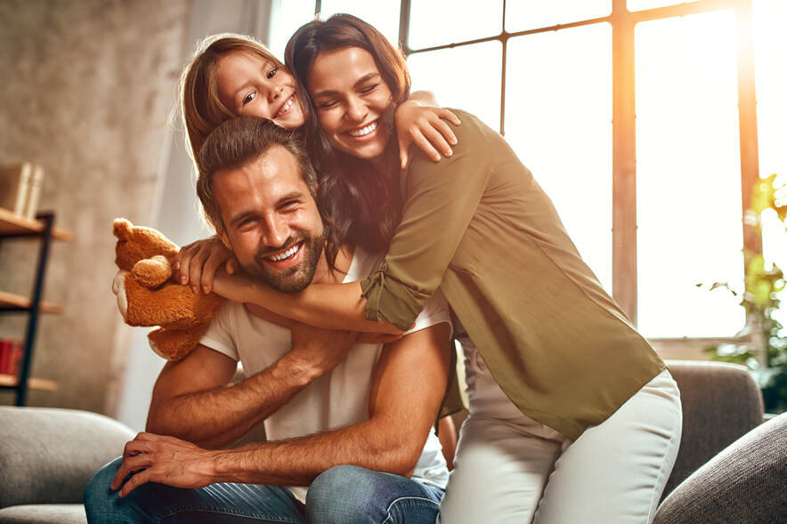 Happy dad and mom with their cute daughter and teddy bear hug and have fun sitting on the sofa in the living room at home.