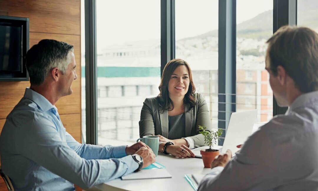 Three business professionals engaged in discussion at a conference table in a modern office setting.