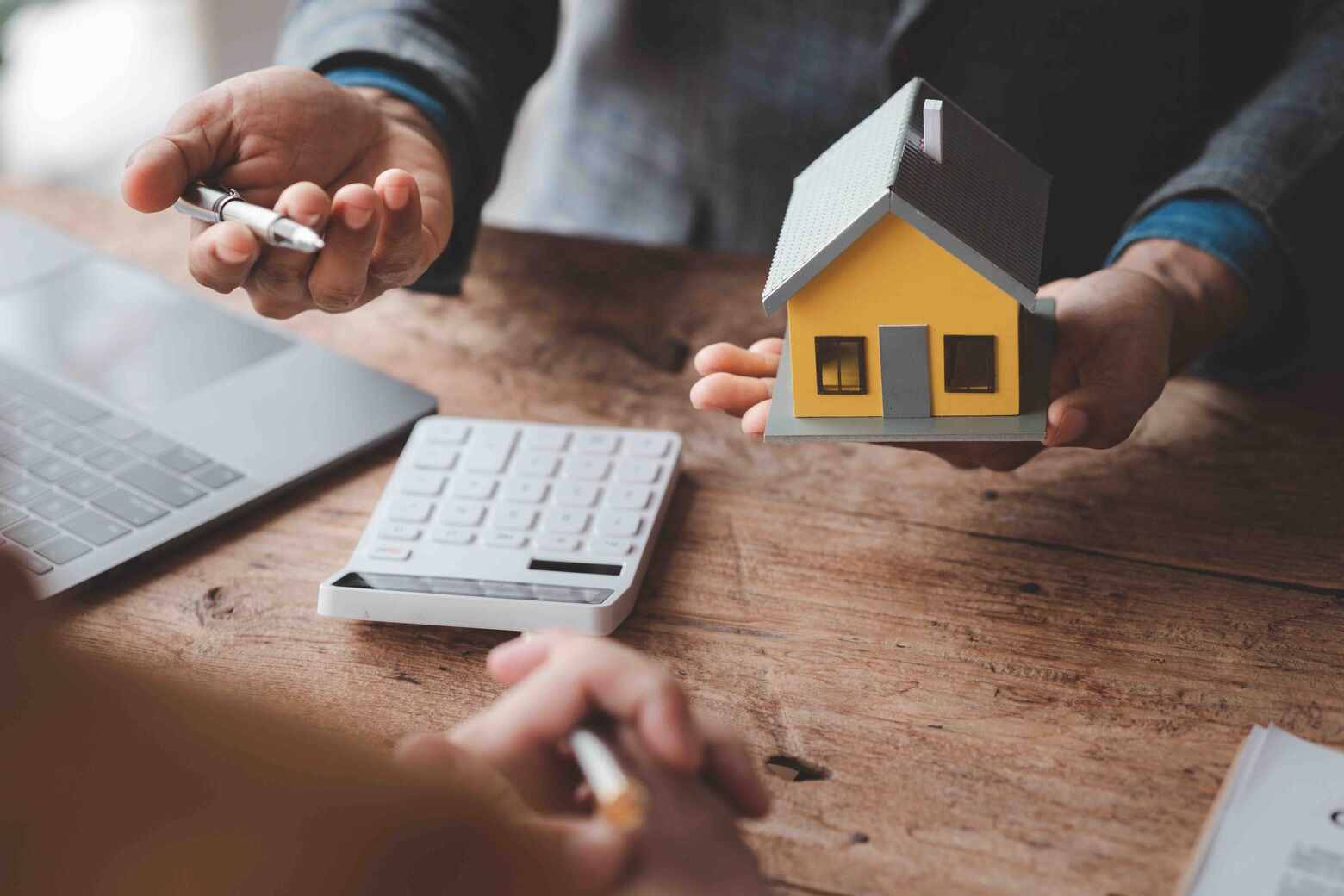 Person holding a model house and a pen, with a calculator and laptop on a wooden table.