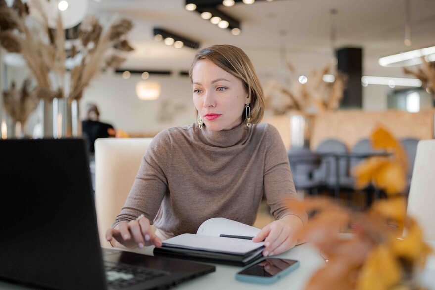 A woman seated at a table, focused on her laptop, with a calm expression in a well-lit room.
