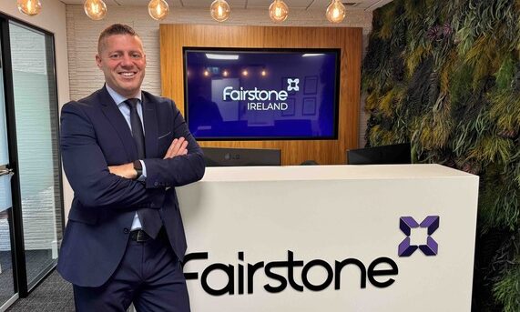 Derek Delany, Fairstone Ireland Managing Director standing at the front desk smiling and with folded arms with Fairstone logo in the background