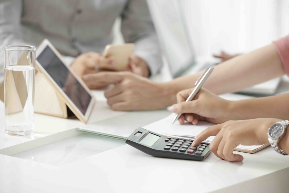 Crop shot of businesswoman using calculating machine and writing down numbers working with colleagues in office