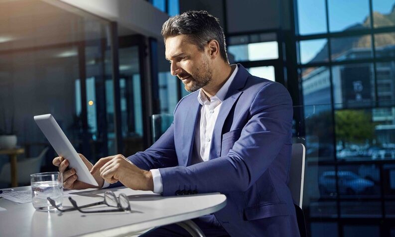 A man in a suit sits at a table, focused on a tablet in front of him, searching information about auto enrolment in Ireland