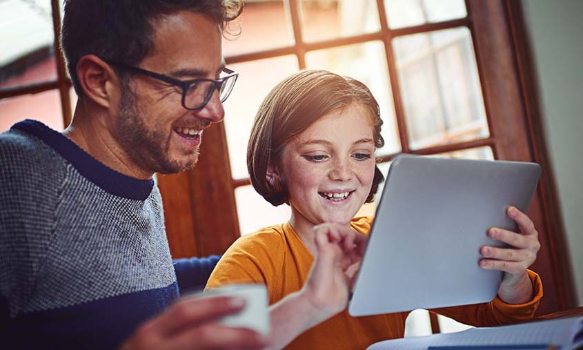 Saving for Education in Ireland - Father and daughter smiling while using a tablet together at home. The man holds a coffee cup, and a notebook lies open on the table. Warm, joyful atmosphere.