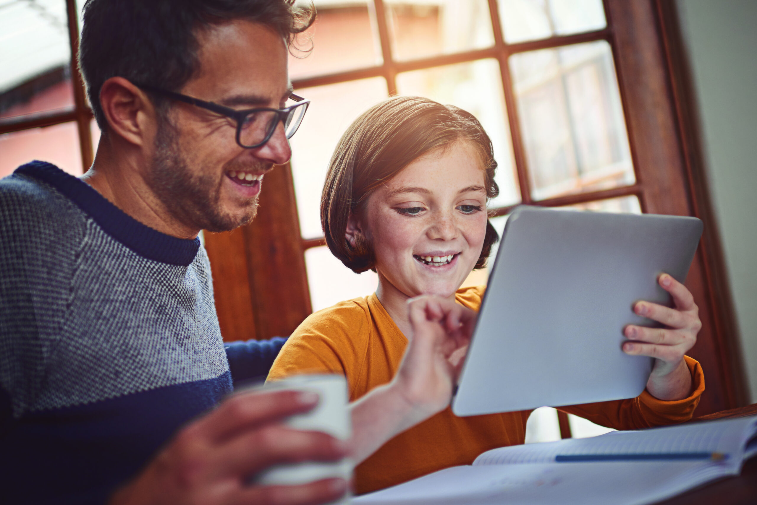 Father and daughter smiling while using a tablet together at home. The man holds a coffee cup, and a notebook lies open on the table. Warm, joyful atmosphere.