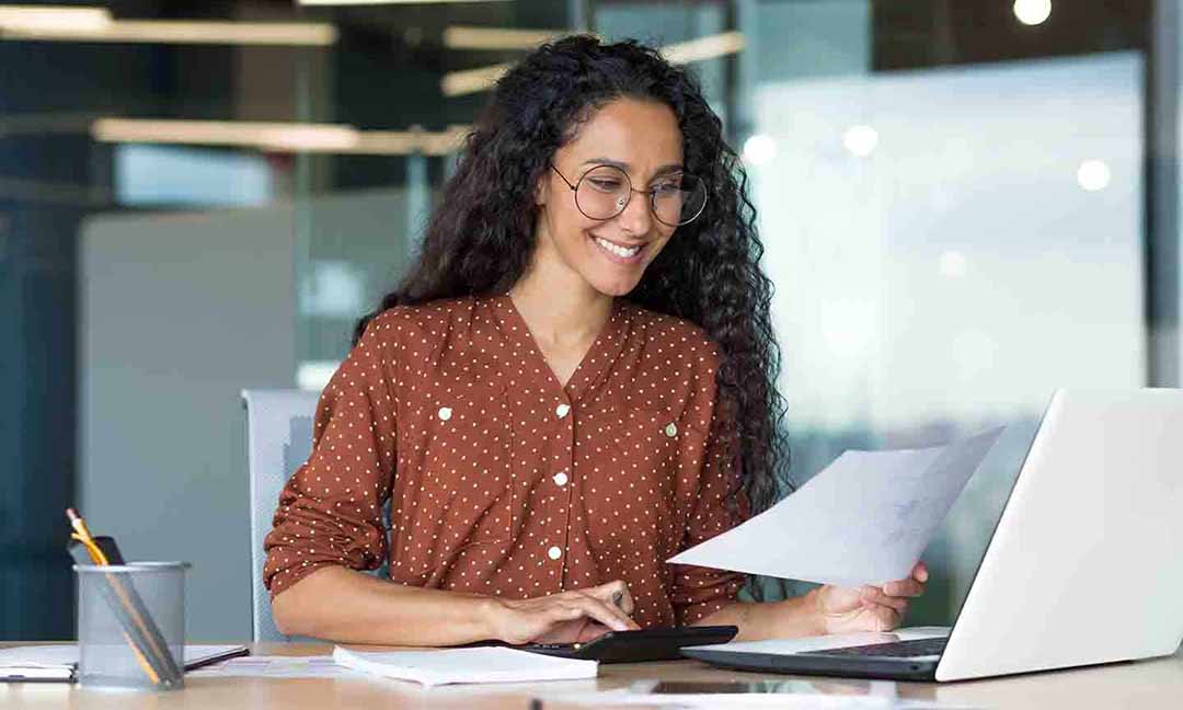 A woman smiles while working on her laptop, conveying a sense of joy and engagement in her task.