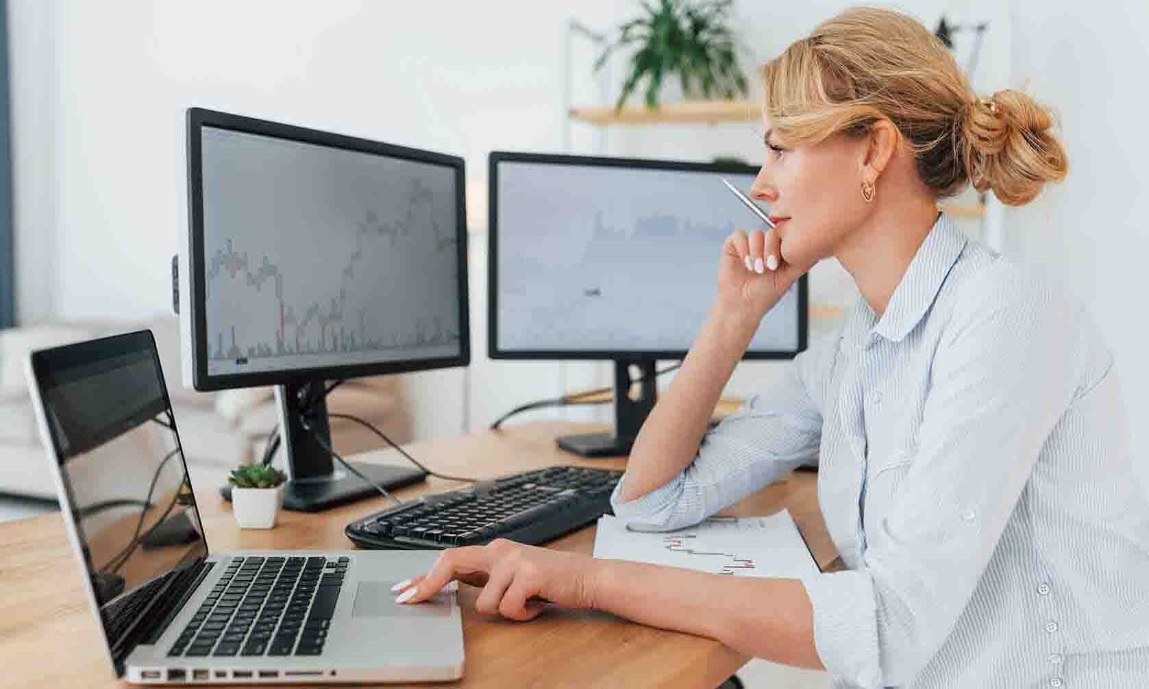 A woman seated at a desk, working with two monitors and a laptop in a modern office setting.